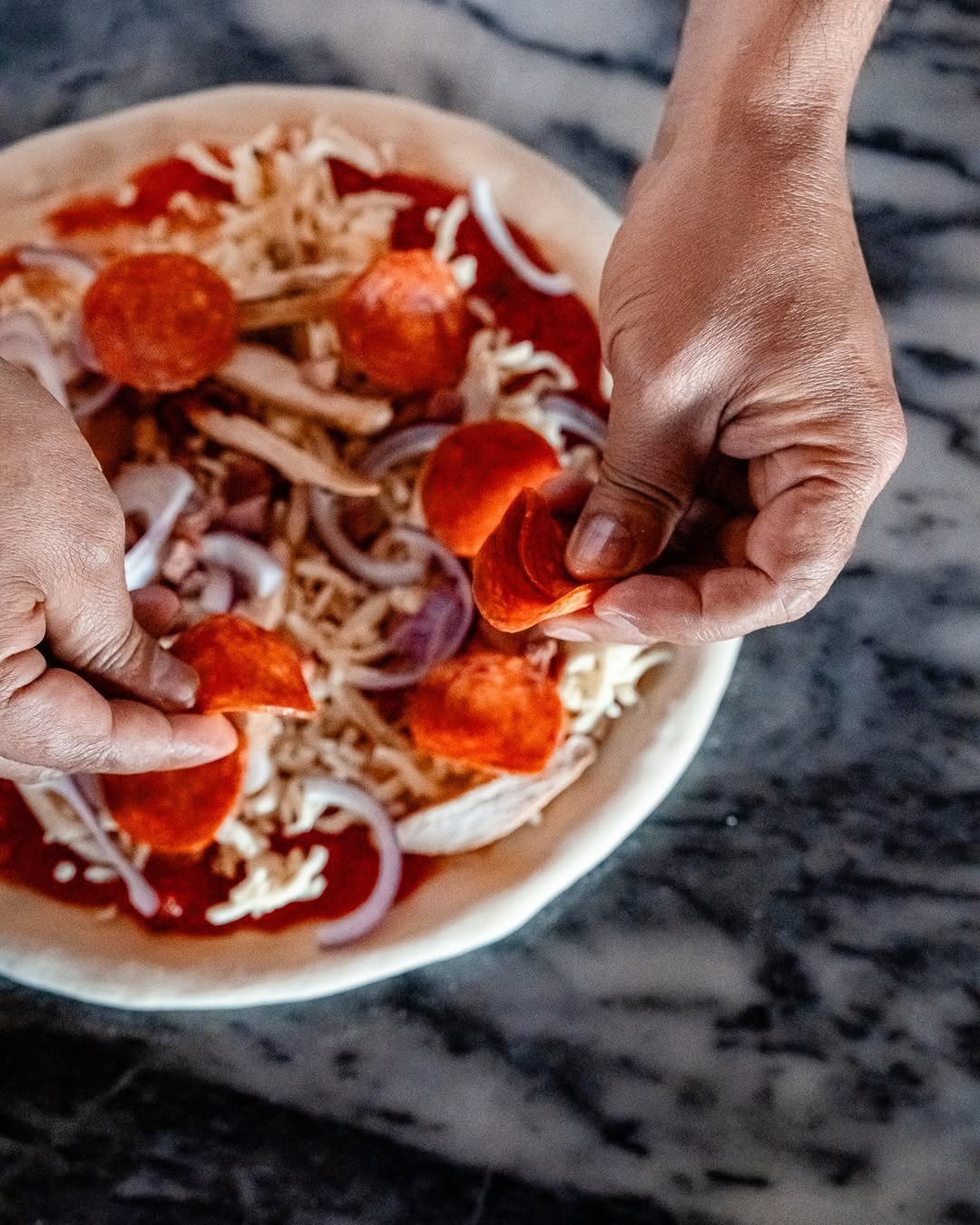 Mãos a espalhar pepperoni e cebola roxa sobre a massa de uma pizza